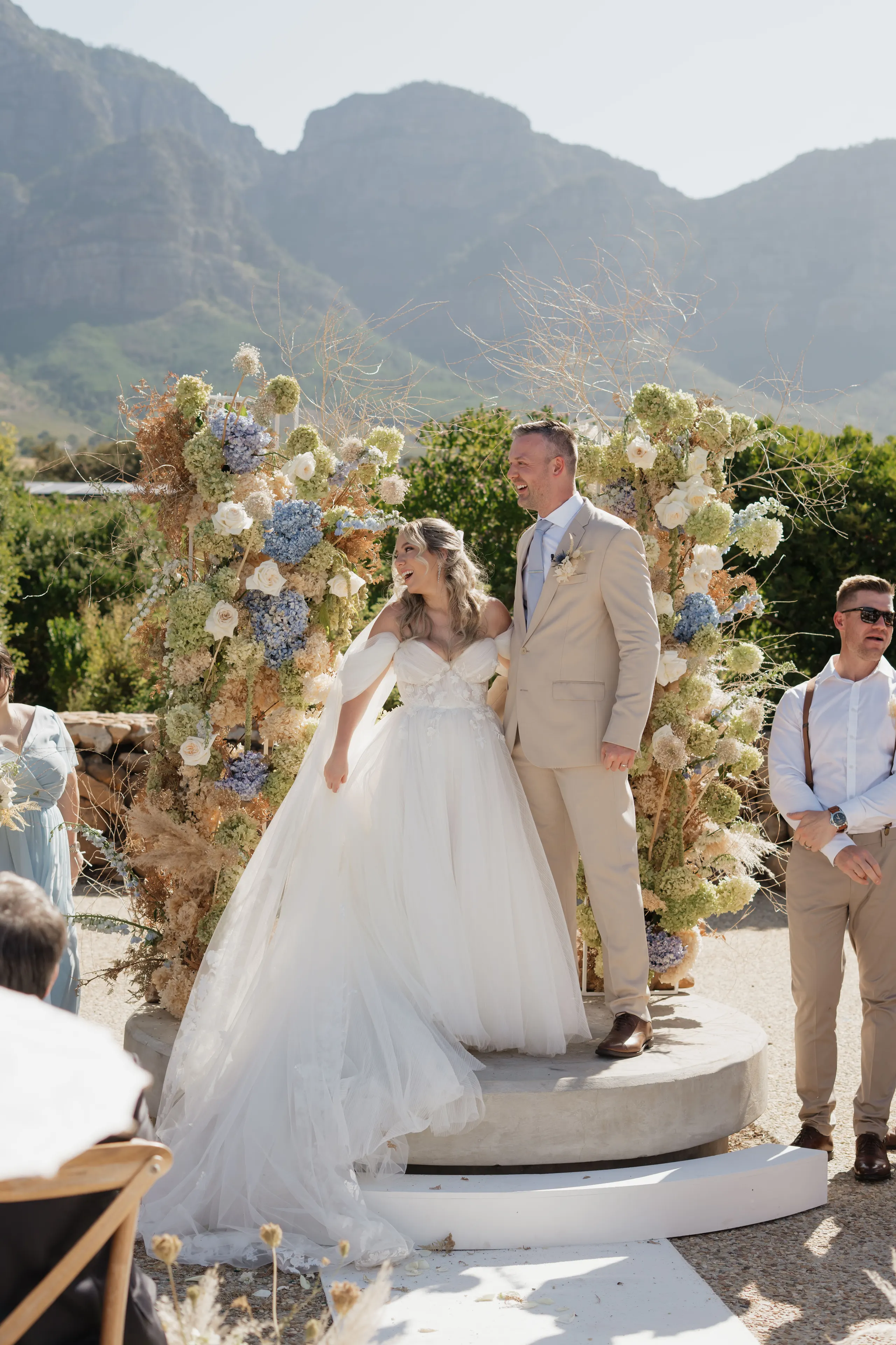 Couple at their ceremony beneath an arch of hydrangeas, guests looking on
