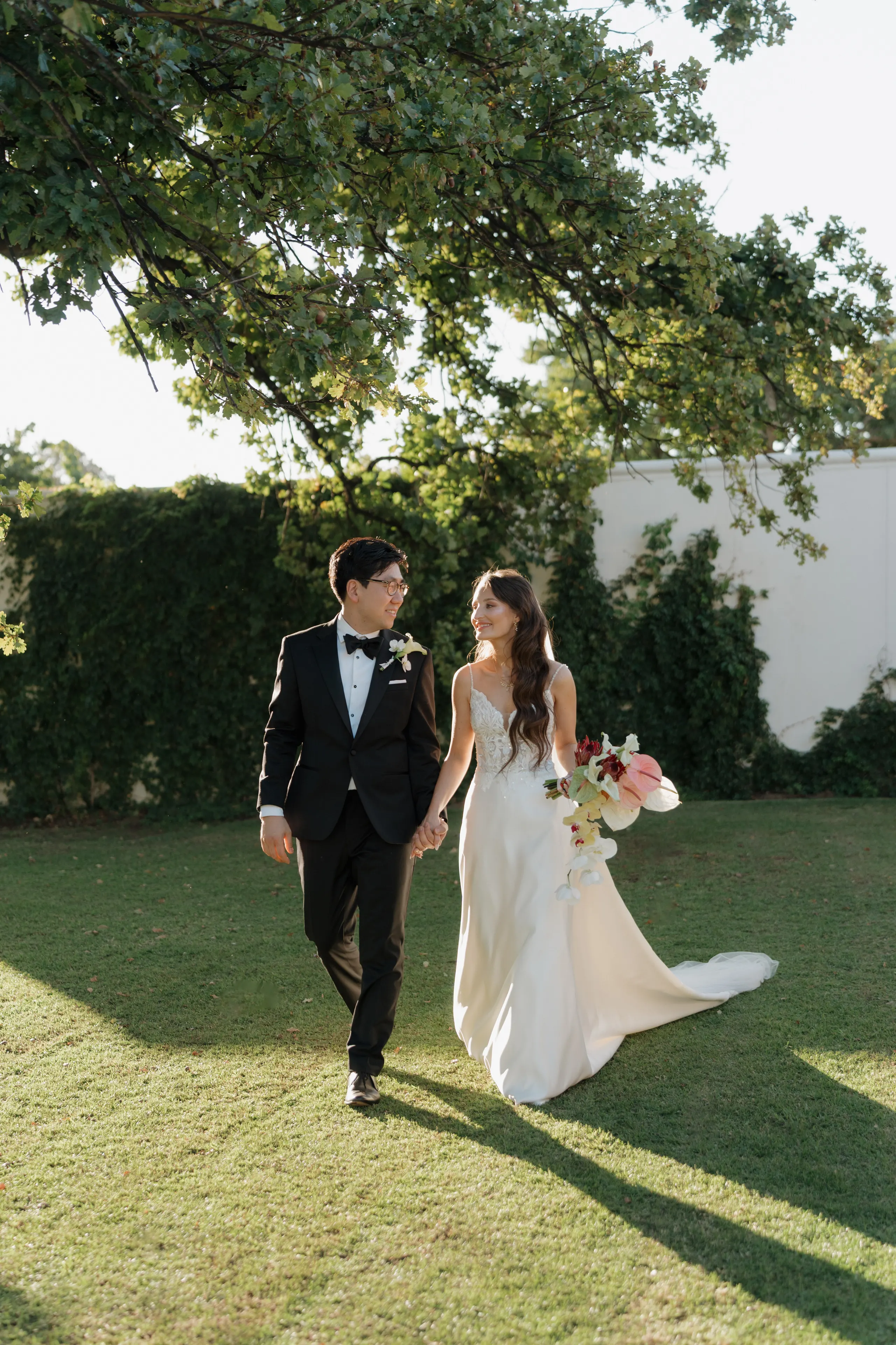Newlyweds walking hand in hand across a garden lawn, bouquet in hand