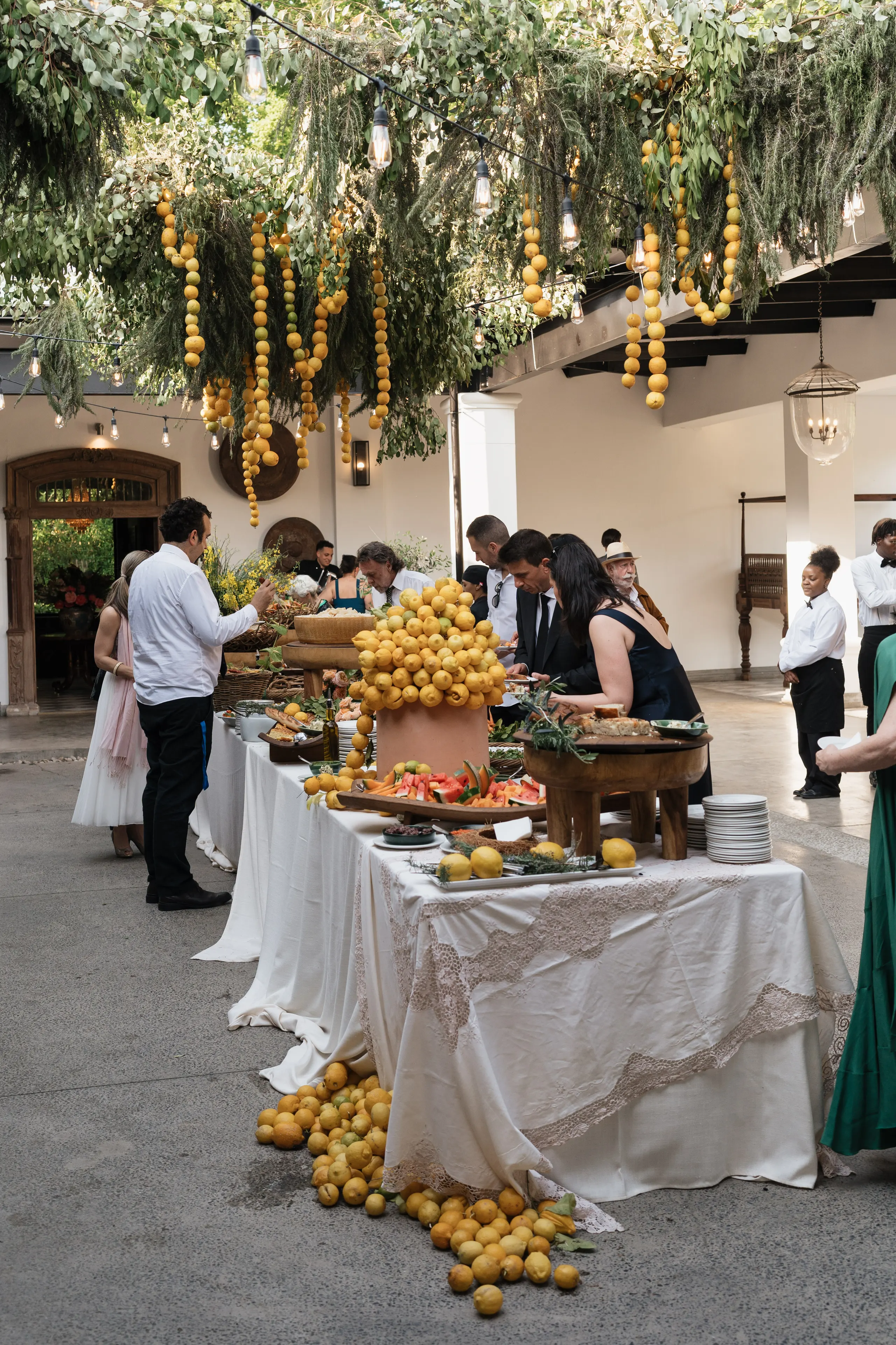 Abundant outdoor feast table piled with lemons beneath hanging citrus, guests gathering