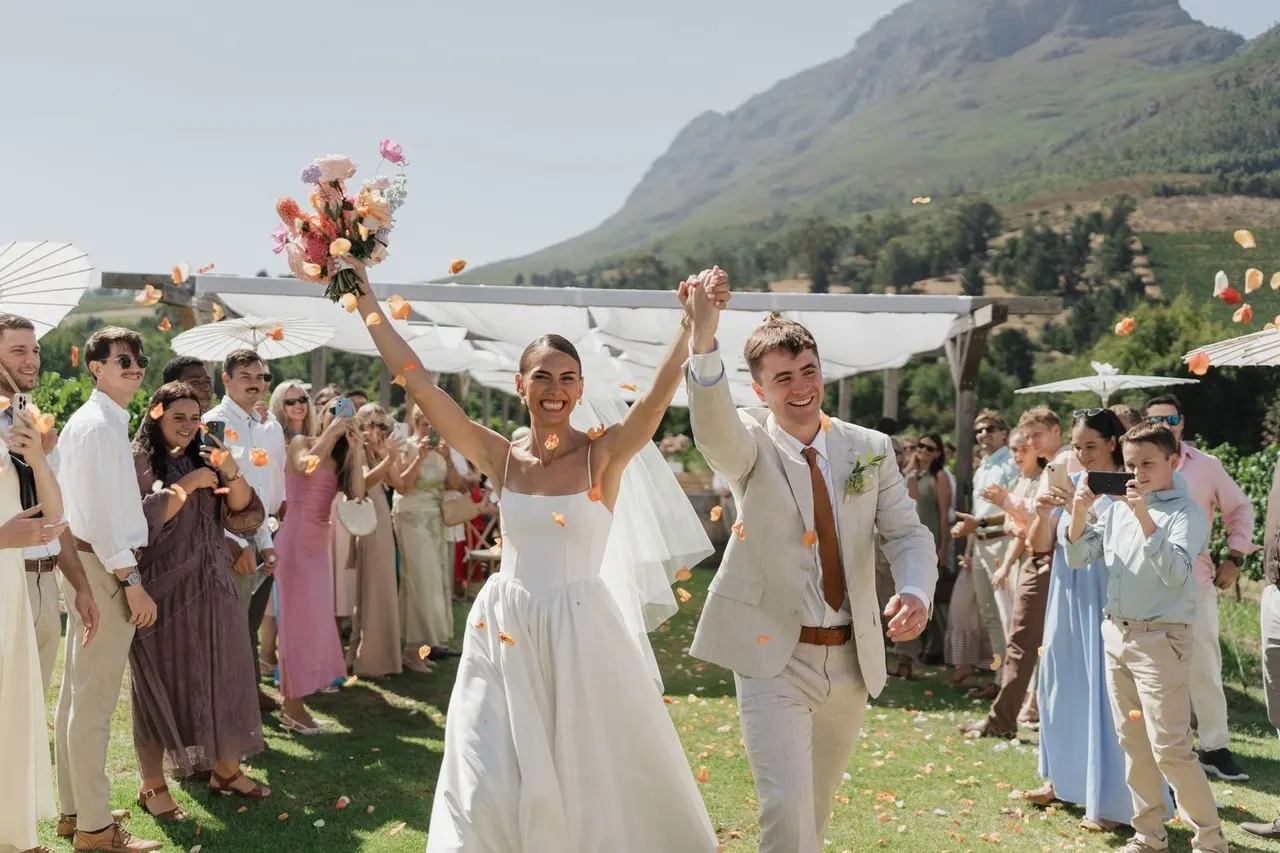 Couple cheering hand in hand beneath ribbons as guests celebrate, mountains behind
