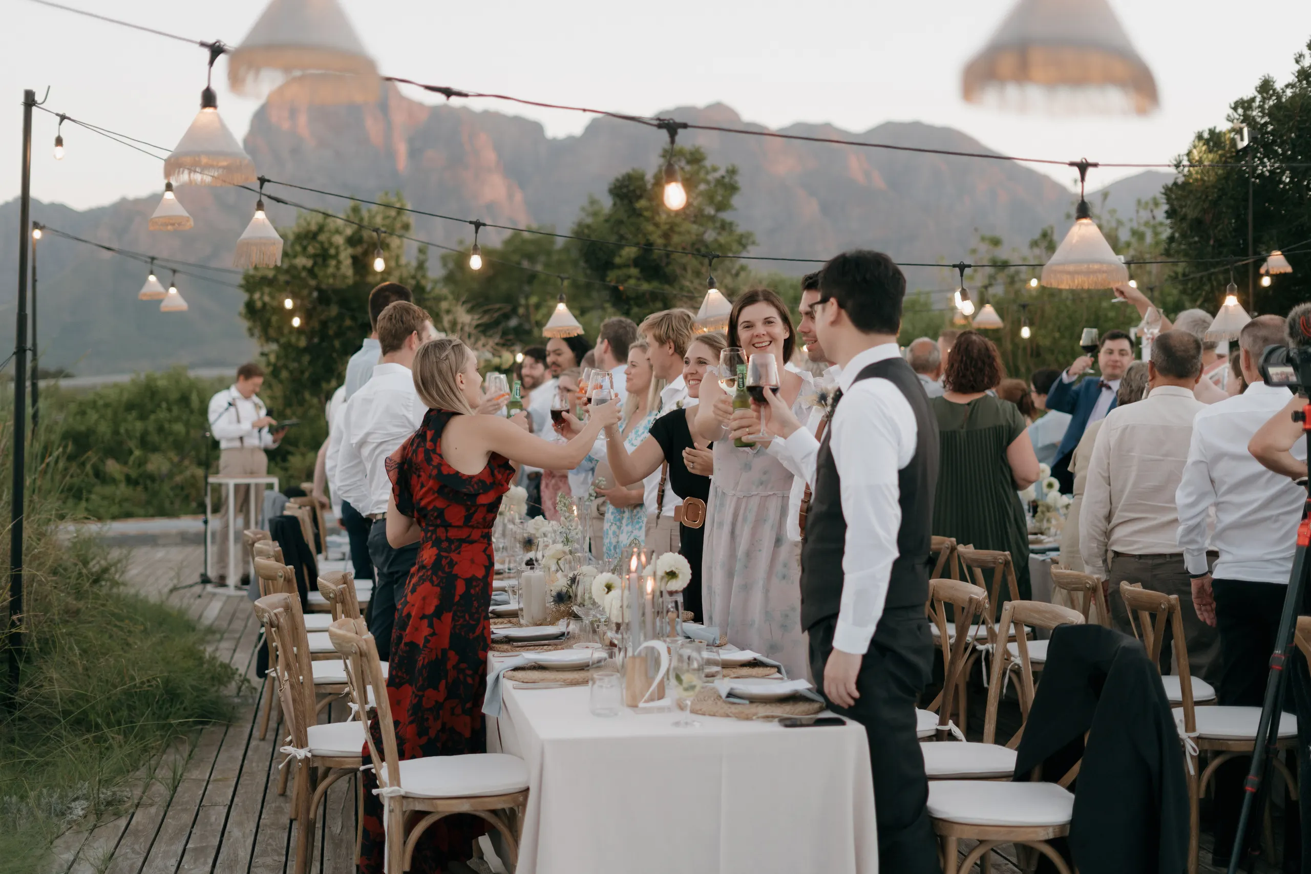Wedding guests raising a toast at long reception tables under string lights, mountains beyond
