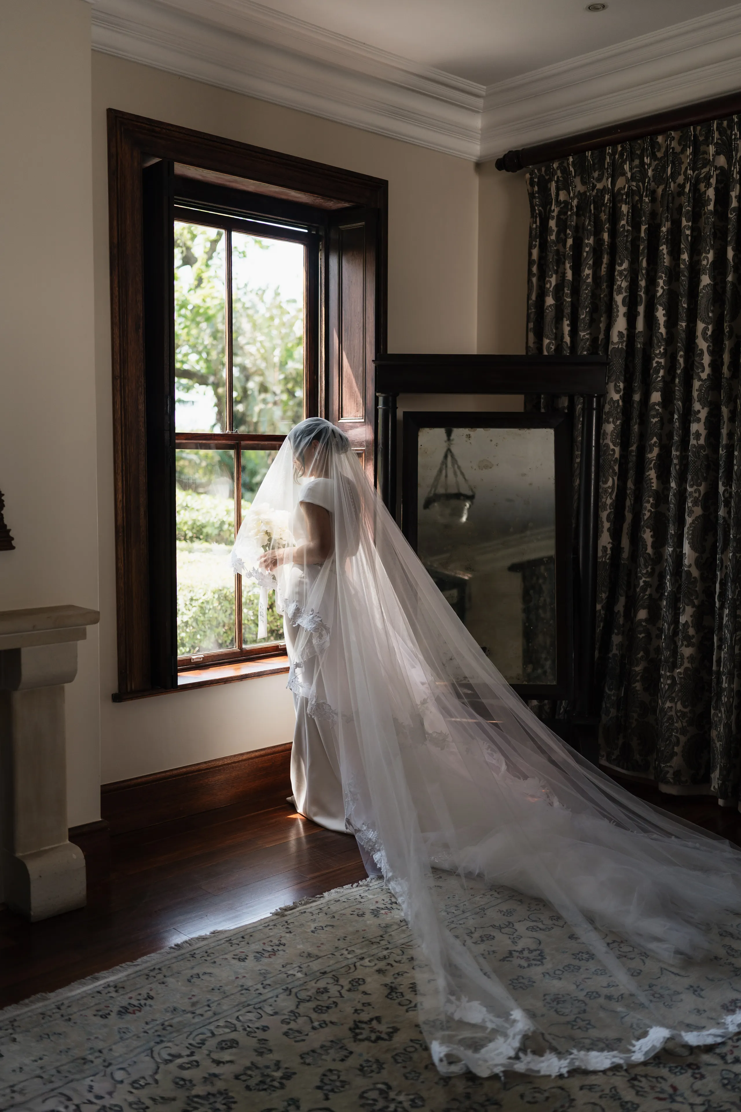 Bride standing at a bright window, her long veil catching the light