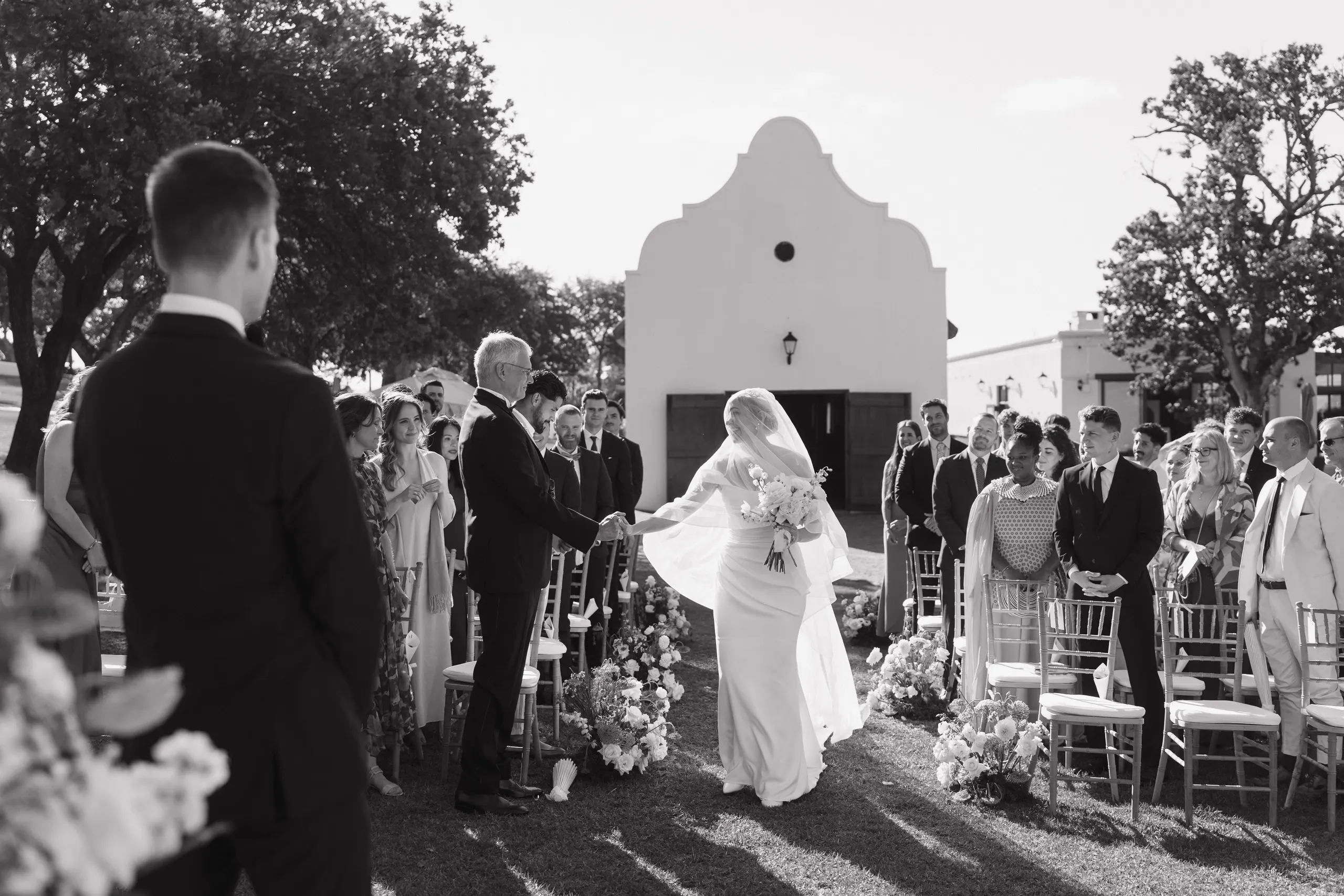 Black-and-white photograph of the bride walking down the outdoor aisle at a Cape Dutch chapel, guests standing